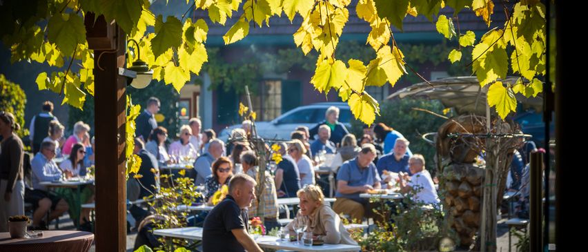 Menschen sitzen an Tischen in einem Außenrestaurant, umgeben von grünen Blättern und einem geparkten Auto. Einige essen, andere plaudern.