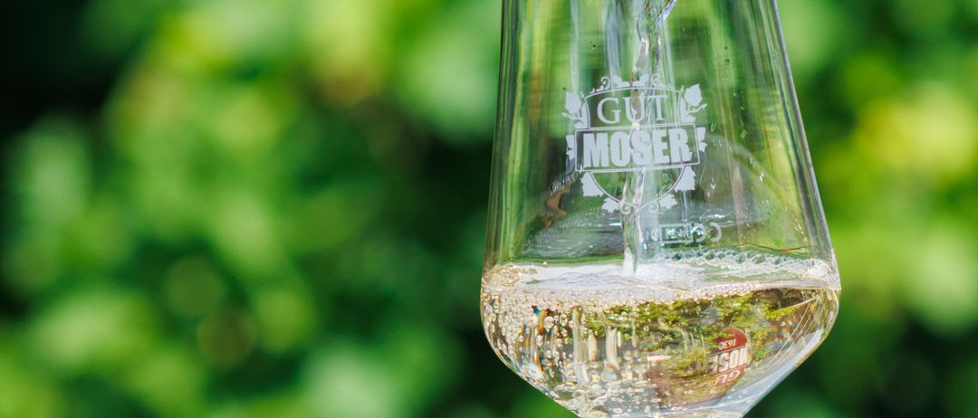 Eine Flasche Gut Moser Wein wird in ein klares Weinglas mit der Aufschrift Gut Moser gegossen. Der Wein ist klar mit Blasen.