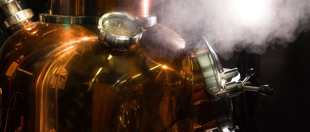 Ein Nahaufnahme einer großen kupferfarbenen Destillationsvorrichtung mit Dampf, der von oben austritt, beleuchtet von einem hellen Licht.