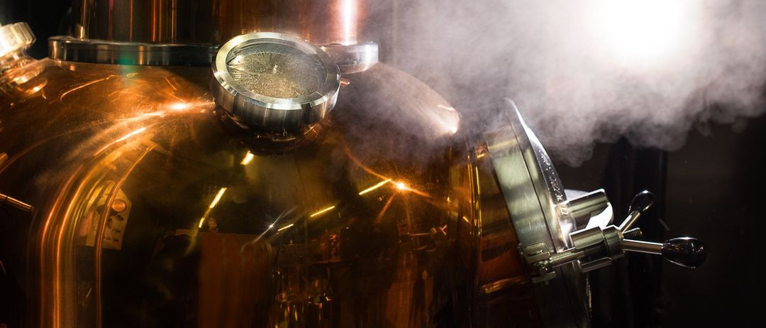 Ein Nahaufnahme eines Kupferdestillierapparats emittiert Dampf. Der Apparat ist poliert und reflektierend.