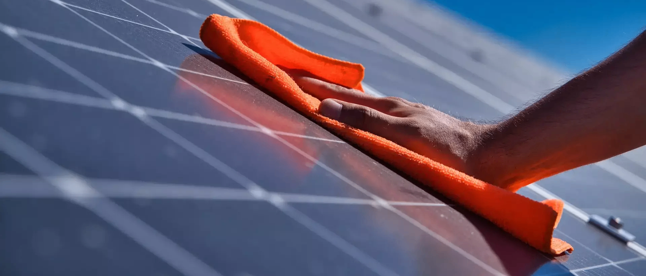 Eine Hand reinigt Solarmodule mit einem orangefarbenen Tuch an einem sonnigen Tag.