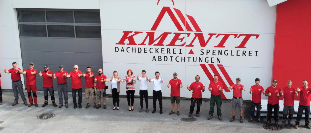 Eine Gruppe von Menschen in roten Hemden und Mützen steht vor einer weißen Wand mit einem roten Logo, das 'KMENTT Dachdeckerei Spengler Abdichtungen' liest. Sie scheinen für ein Foto zu posieren.
