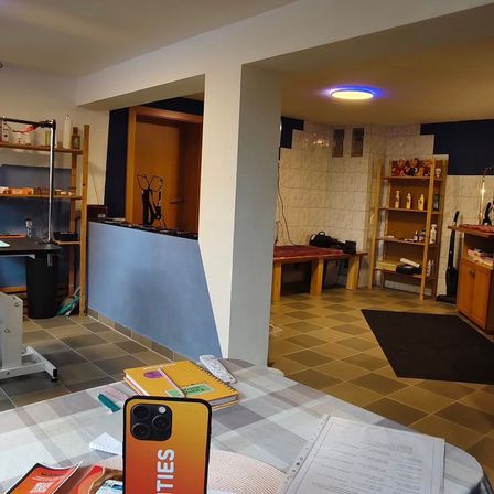 Ein Zimmer mit einem Schreibtisch, Büchern und einem Smartphone mit dem Wort 'CITIES' darauf. Regale, ein Tisch und eine Bank sind ebenfalls vorhanden. Eine Deckenlampe ist beleuchtet.