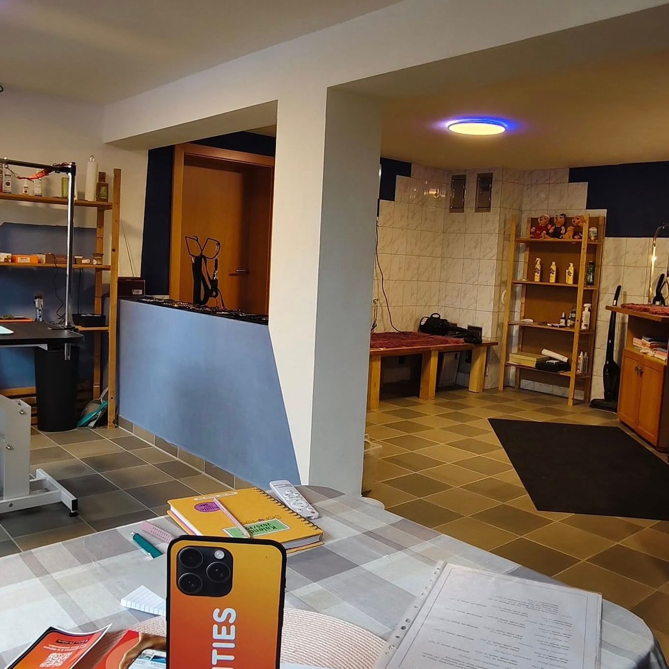 Ein Zimmer mit einem Schreibtisch, Büchern und einem Smartphone mit dem Wort 'CITIES' darauf. Regale, ein Tisch und eine Bank sind ebenfalls vorhanden. Eine Deckenlampe ist beleuchtet.