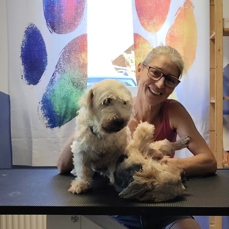Eine Frau hält zwei Hunde in einem Raum mit einem farbenfrohen Pfotenabdruck im Hintergrund. Die Frau lächelt, während sie auf einem Tisch sitzt, auf dem die Hunde sitzen.