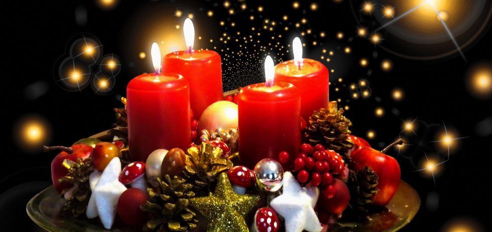 Vier rote Kerzen brennen auf einem goldenen Teller, umgeben von Ornamenten und Tannenzapfen. Der schwarze Hintergrund hat gelbe Lichter in der Ferne.