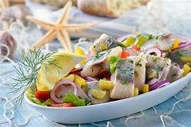 Ein weißer Teller auf einem blauen Tisch enthält ein Gericht aus Fisch, Gemüse und Kräutern. Im Hintergrund sind Seesterne und Brot zu sehen.