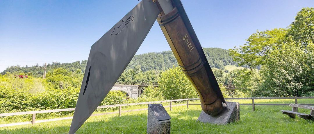 A large outdoor sculpture of a knife stands in a grassy field. The blade is silver, and the handle is brown.