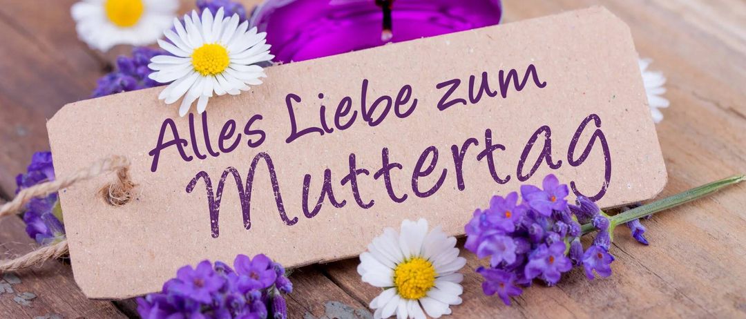 A candle with a note saying Alles Liebe zum Muttertag is surrounded by flowers on a wooden surface.