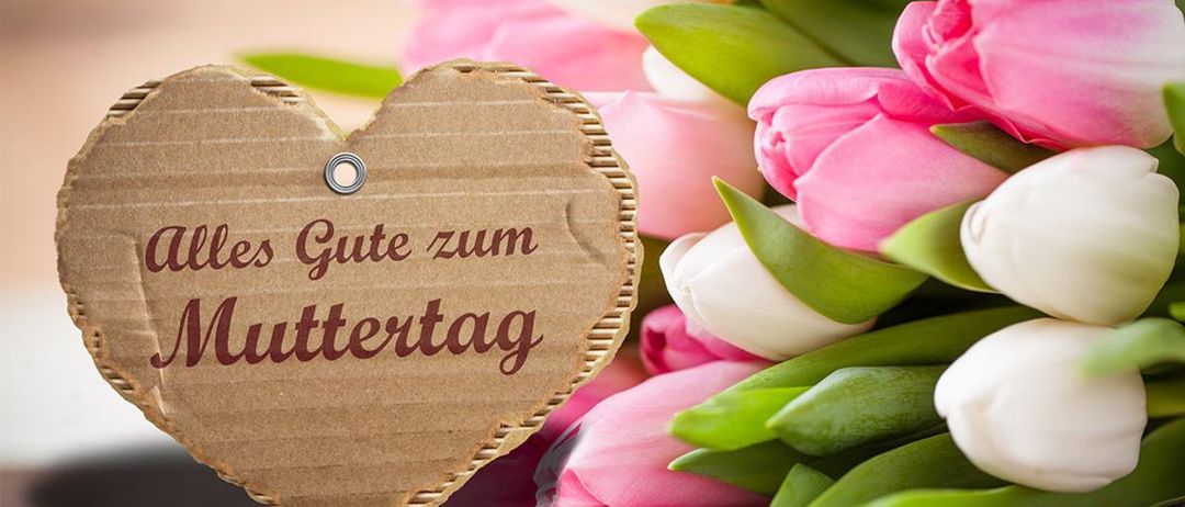 Ein herzförmiger Pappaufkleber mit 'Alles Gute zum Muttertag' in Rot. Ein Strauß rosa und weißer Tulpen im Hintergrund.