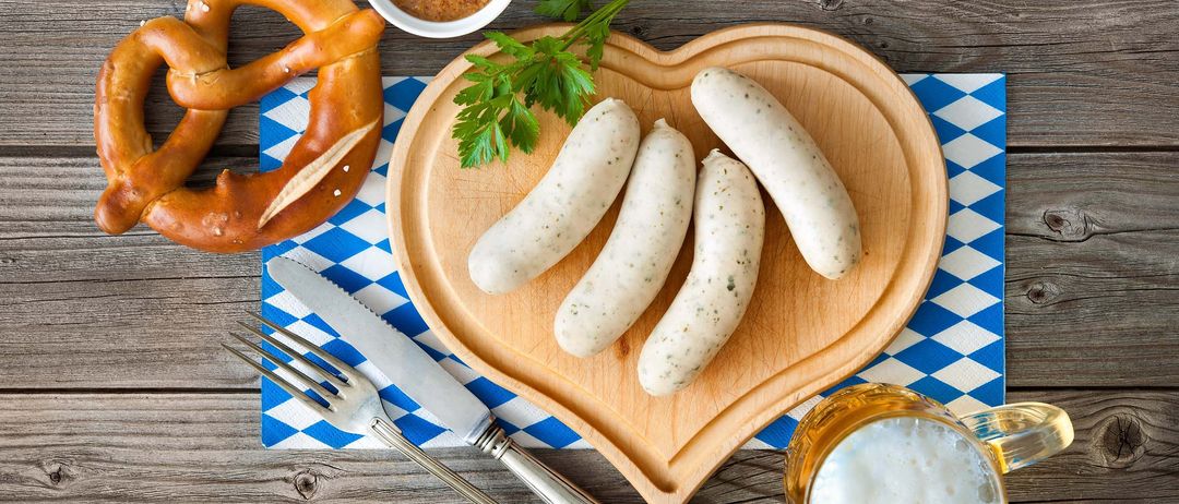 Vier Würste auf einem herzförmigen Holzschneidebrett, garniert mit Petersilie, mit einer Schüssel Senf, einem Brezel und einem Glas Bier auf einer blau-weiß karierten Tischdecke.