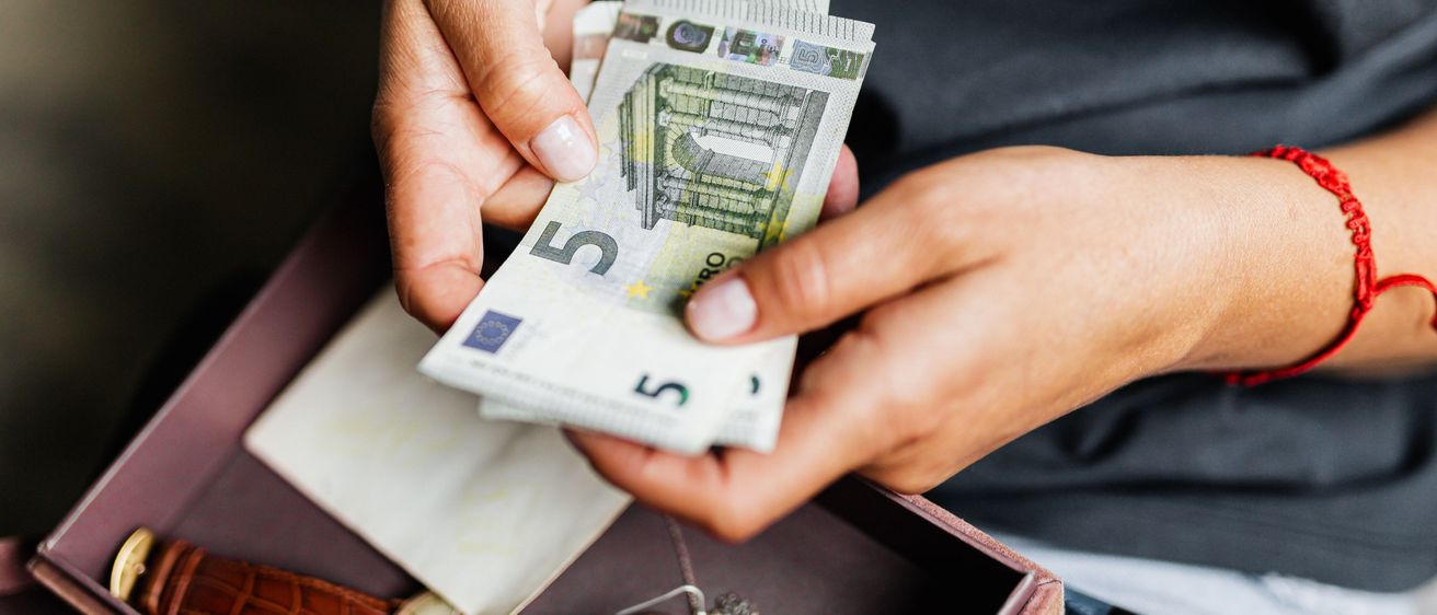 Eine Person hält einige Euro-Banknoten in der Hand, während sie auf einem Sofa sitzt, neben einer Schachtel und einer Uhr.