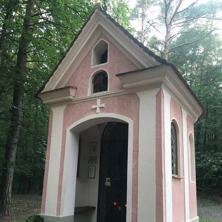 Eine kleine, rosafarbene Kapelle mit einem Kreuz oben, einem Glasfenster und einer Bank in einem Wald. Die Tür ist geschlossen, und eine Kerze ist im Inneren angezündet.