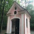 Eine kleine, rosafarbene Kapelle mit einem Kreuz oben, einem Glasfenster und einer Bank in einem Wald. Die Tür ist geschlossen, und eine Kerze ist im Inneren angezündet.