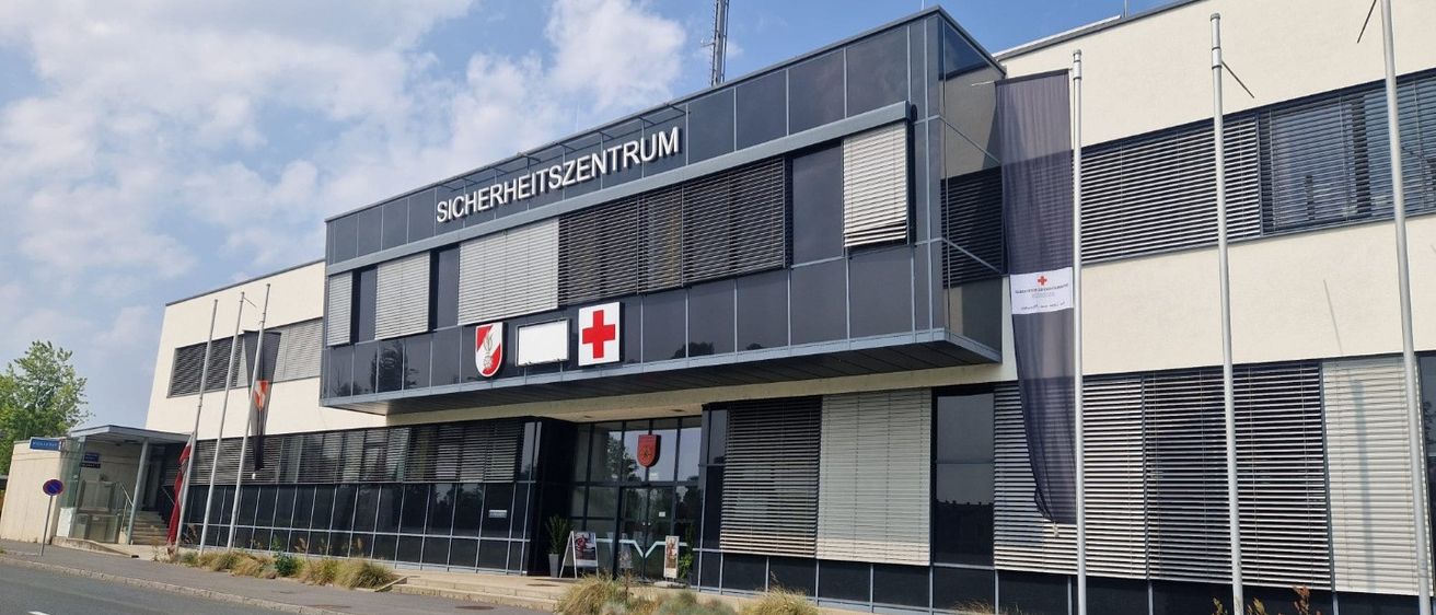 Ein modernes Gebäude mit dem Wort 'Sicherheitszentrum' darauf, einem roten Kreuz-Schild und einem Fahnenmast. Es hat viele Fenster und einen Weg davor.