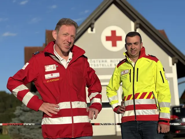 Zwei Männer in roten und gelben Sicherheitsjacken stehen vor einem Gebäude mit einem roten Kreuzschild an der Wand.
