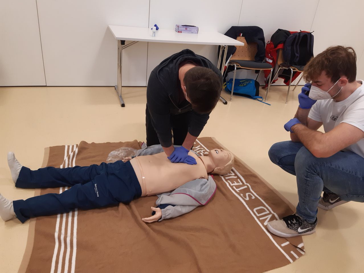 Zwei Personen führen Herz-Lungen-Wiederbelebung an einer Puppe auf einem braunen Teppich durch. Sie tragen Masken und Handschuhe. Ein Tisch und Stühle sind im Hintergrund.