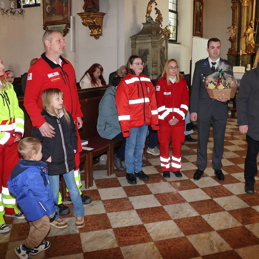 In einer Kirche stehen mehrere Personen in roten und weißen Sicherheitsjacken, während andere auf Bänken sitzen. Ein Mann in Uniform hält einen Korb.