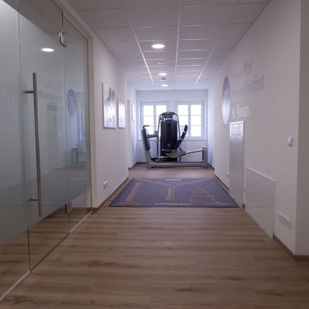 Ein leerer Flur mit einer Glastür und einer Glaswand, die zu einem anderen Raum führt. Ein grauer Teppich mit Logo liegt auf dem Boden. In der Mitte des Flurs befindet sich ein Fitnessgerät. An der Wand hängen Bilderrahmen und ein Schild.