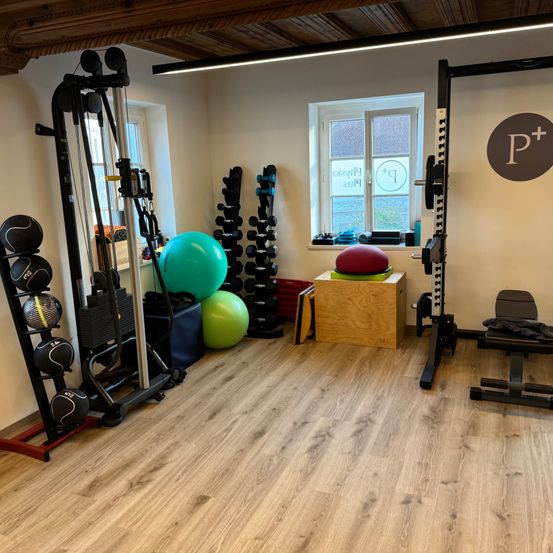 Ein Fitnessstudio mit Holzböden und weißen Wänden. Verschiedene Trainingsgeräte und Gewichte sind im Raum verteilt. Ein Holzkasten hält Übungsbälle. Ein Fenster an der Wand zeigt das Außen.