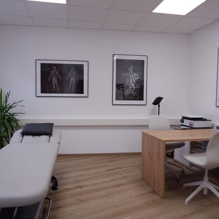 Ein Büro mit Holzböden und weißen Wänden. Zwei gerahmte Bilder hängen an der Wand. Ein Schreibtisch mit Stuhl ist im Raum. Ein Massagetisch mit Kissen ist auf der linken Seite.