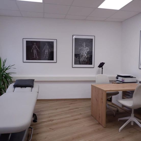 Ein Büro mit Holzböden und weißen Wänden. Zwei gerahmte Bilder hängen an der Wand. Ein Schreibtisch mit Stuhl ist im Raum. Ein Massagetisch mit Kissen ist auf der linken Seite.