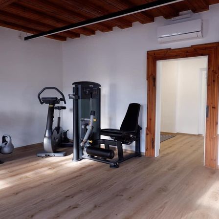 Ein leerer Fitnessraum mit Holzboden, weißen Wänden und verschiedenen Trainingsgeräten, darunter ein Laufband, eine Bank und eine Kettlebell.