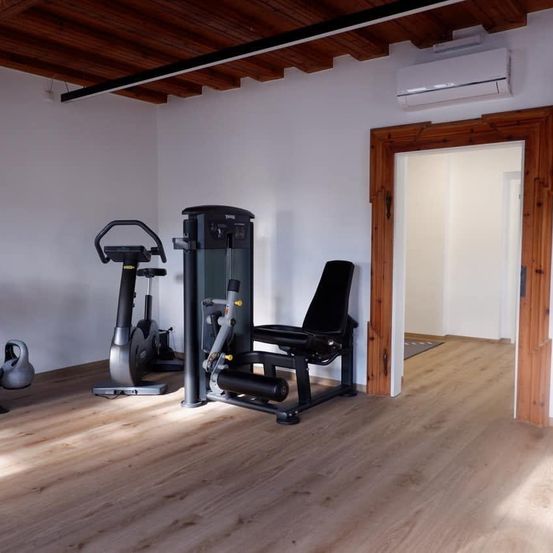 Ein leerer Fitnessraum mit Holzboden, weißen Wänden und verschiedenen Trainingsgeräten, darunter ein Laufband, eine Bank und eine Kettlebell.