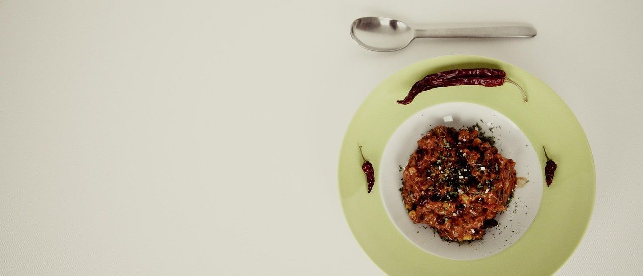 Ein Gericht mit roten Chilis und Kräutern auf einem grünen Teller mit einem Löffel. Das Gericht wird auf einer weißen Oberfläche serviert.