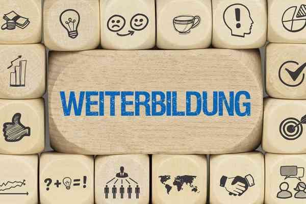 Ein Holzbrett mit dem Wort Weiterbildung darauf, umgeben von verschiedenen Symbolen und Icons wie Lichter, Gesichter und eine Tasse.