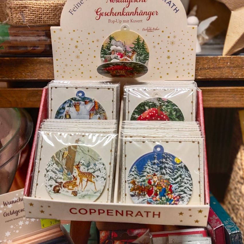 Eine Anzeige von Weihnachtsgeschenk-Anhängern von Coppenrath, mit Winterszenen, präsentiert in einer Box mit einem Pop-Up-Fenster.