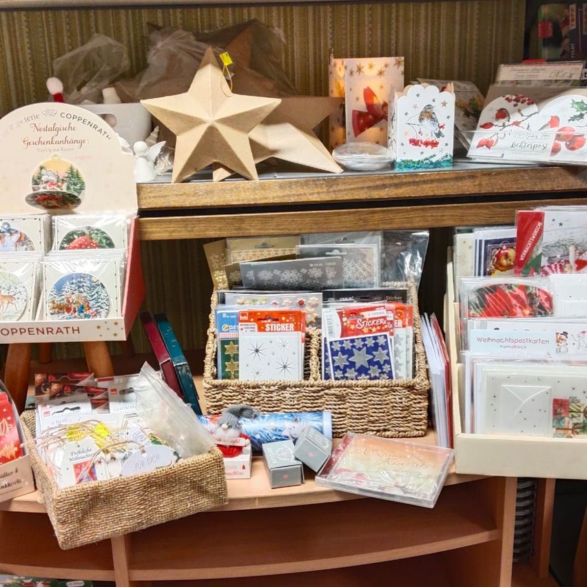 Ein Holzregal gefüllt mit verschiedenen Weihnachtsartikeln. Es zeigt ein Sternornament, Grußkarten, Aufkleber und Dekorationsartikel. Die Karten sind in Kisten und Körben angeordnet, und einige haben Weihnachtsmotive.