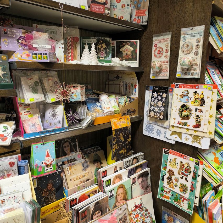 Ein hölzerne Regal mit verschiedenen Weihnachtsdekorationen, Karten und Aufklebern gefüllt. Das Regal hat mehrere Ebenen mit unterschiedlichen Artikeln wie Weihnachtskarten, Aufklebern und Dekorationsgegenständen. Einige Artikel sind ordentlich organisiert, während andere in verstreuten Kisten sind.