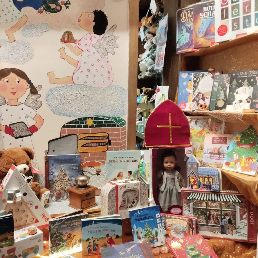 Eine weihnachtliche Ausstellung zeigt eine Wandmalerei von Engeln, eine Puppe, Bücher, einen Teddybär, einen Nussknacker und eine Kaffeemühle. Ein Holzregal hält verschiedene Bücher und Spielzeug.