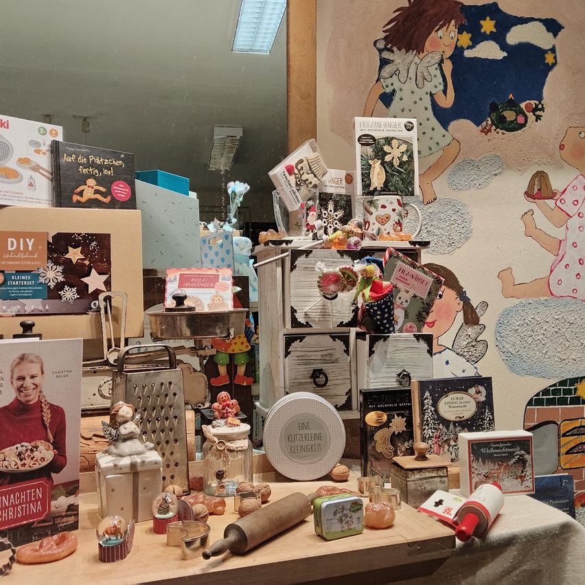 Ein Tisch zeigt verschiedene Küchenutensilien, darunter ein Nudelholz, eine Messbecher und eine Reihe von Weihnachtsdekorationen. Im Hintergrund ist ein Wandbild mit Kindern, Sternen und Wolken zu sehen.