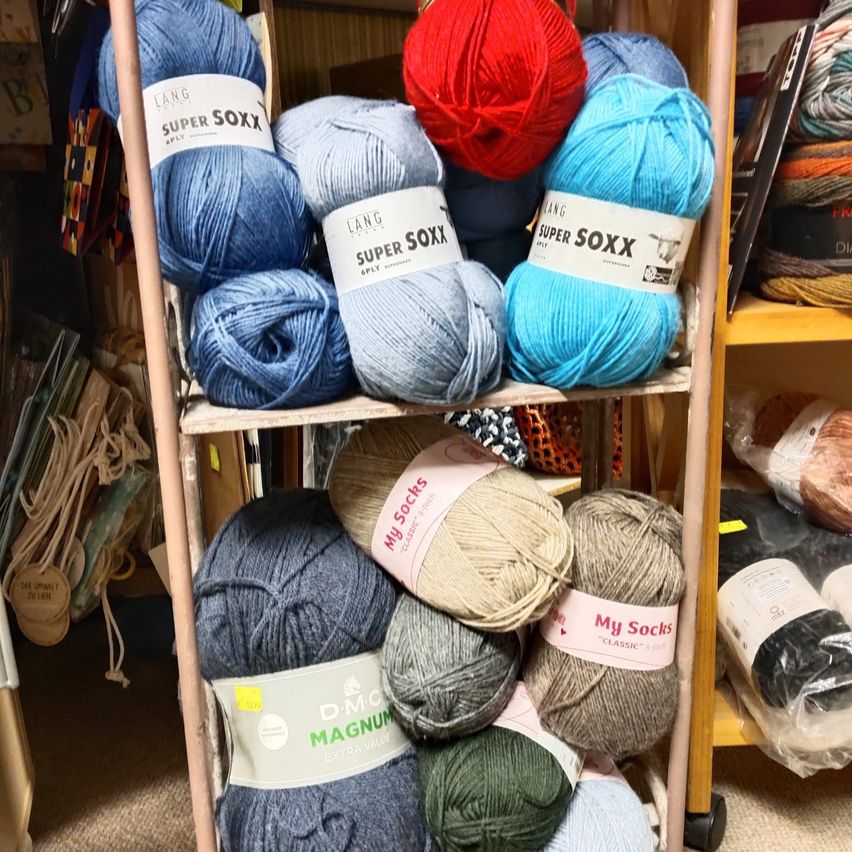 Ein Holzregal zeigt verschiedene Garnrollen, darunter Super Soxx, My Socks und Magnum. Die Garnrollen kommen in verschiedenen Farben, darunter Blau, Rot und Beige. Im Hintergrund sind zusätzliche Garne und Objekte zu sehen.