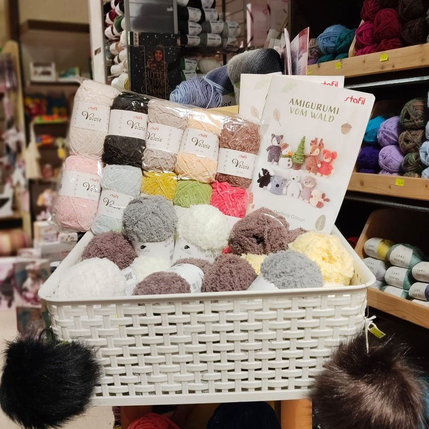 Ein weißer Korb voller verschiedenfarbiger Wollknäuel, einige in Plastik eingewickelt. Ein Buch mit dem Titel Amigurumi vom Wald liegt oben auf der Wolle. Im Hintergrund sind Regale mit Wollknäueln zu sehen.
