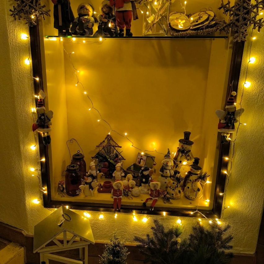 Eine gelb gestrichene Vitrine mit Weihnachtsfiguren und dekorativen Lichtern. Ein kleines hausähnliches Gebilde mit Schlüsselloch befindet sich unten.