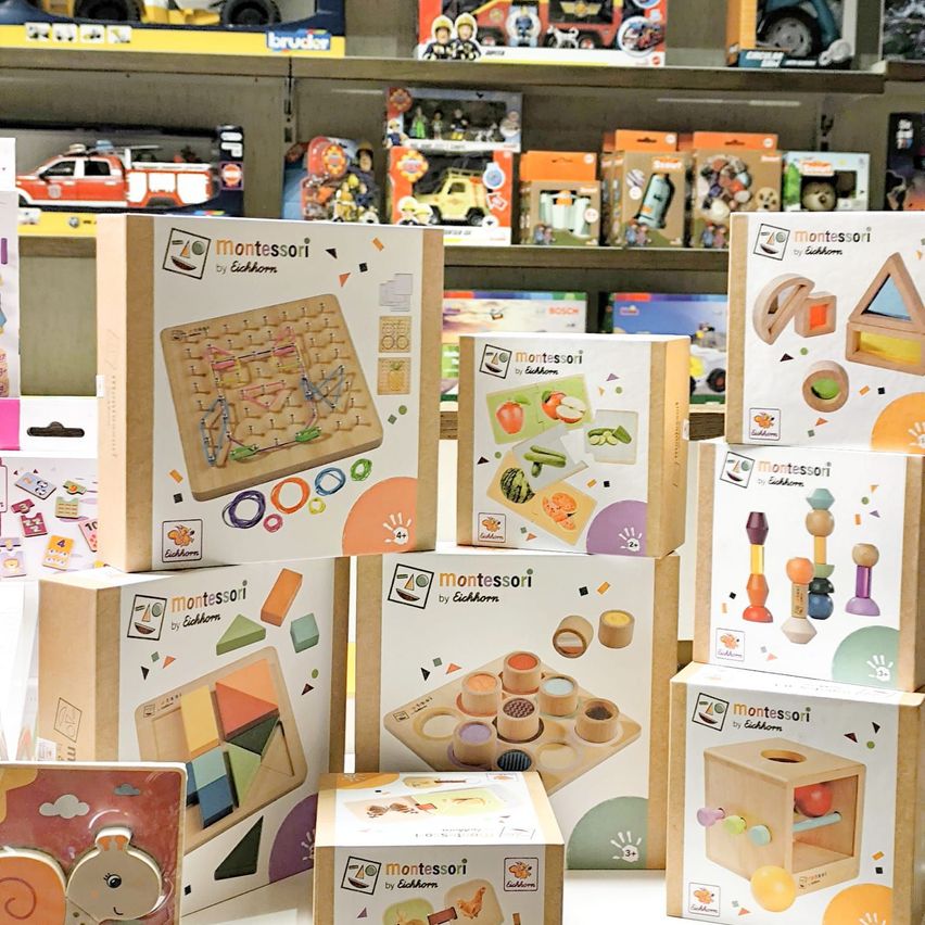 Eine Vielzahl von Montessori-Lernspielzeug ist auf einem Regal ausgestellt. Die Spielzeuge umfassen Puzzles, Bauklötze und andere interaktive Lernwerkzeuge. Jedes Spielzeugkarton hat das Montessori-Logo und die Eichhorn-Marke.