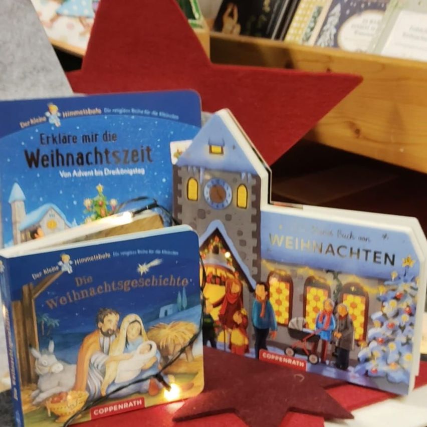 Drei weihnachtlich gestaltete Bücher und ein kleines hausförmiges Objekt mit Licht sind auf einem Tisch ausgestellt. Die Bücher umfassen 'Erklare mir die Weihnachtszeit' und 'Die Weihnachtsgeschichte'. Das Haus hat ein beleuchtetes Fenster und eine Tür.