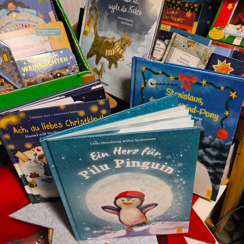 Verschiedene Kinderbücher sind ausgestellt, wobei ein Buch mit dem Titel 'Ein Herz für Pilu Penguin' im Vordergrund einen Pinguin auf dem Cover zeigt. Andere Bücher sind 'Ach, du liebes Christkind' und 'Weihnachten achtslieder'.