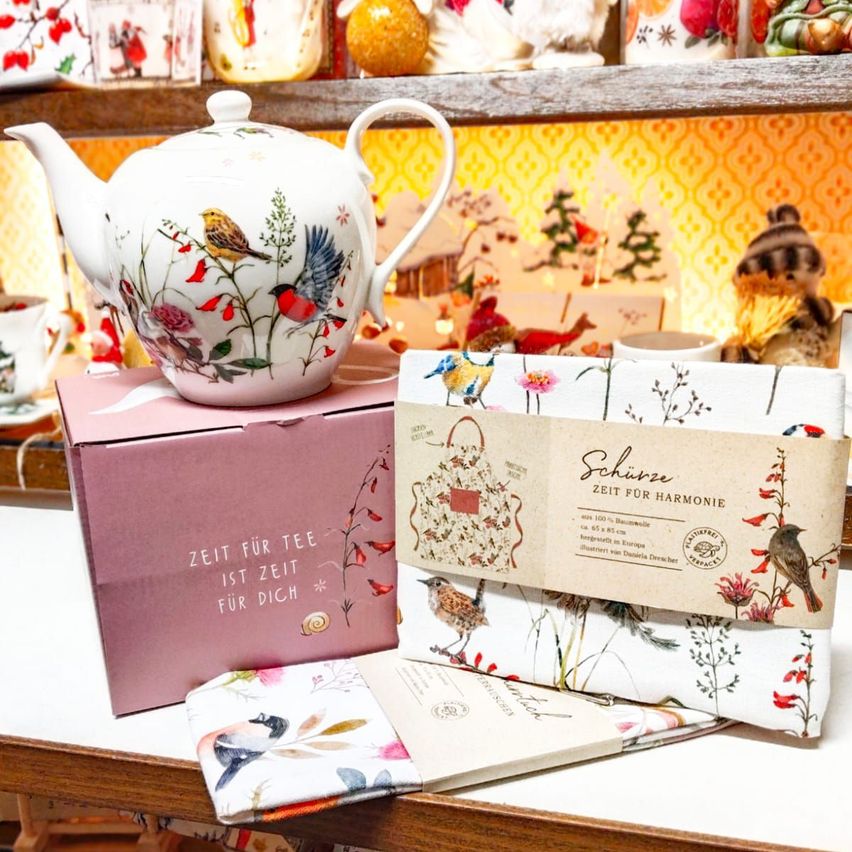 Ein Teekanne mit Vogeldesigns steht auf einer rosa Box mit der Aufschrift 'Zeit für Tee ist Zeit für Dich'. Daneben befindet sich eine Packung mit Blumendesign und der Aufschrift 'Schonzeit für Harmonie'. Darunter sind gefaltete Servietten mit ähnlichen Designs. Dahinter befindet sich ein Regal mit verschiedenen Gegenständen.
