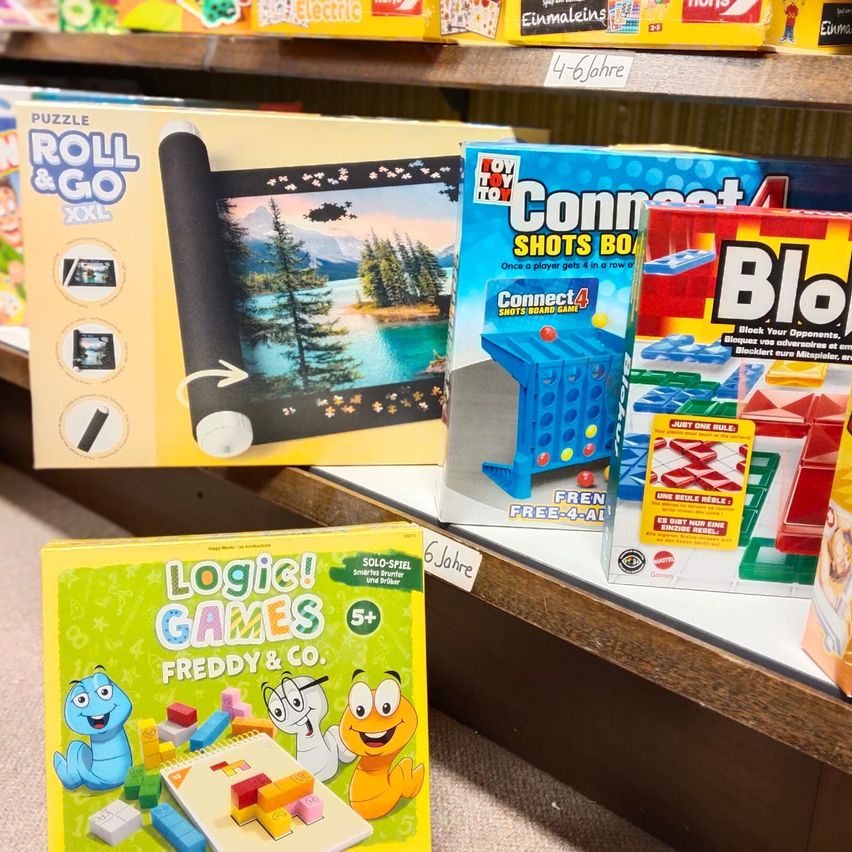 Ein Holzregal zeigt verschiedene Brettspiele, darunter 'Puzzle Roll & Go XXL', 'Connect 4 Shots Board Game' und 'Logic Games Freddy & Co.'. Altersempfehlungen sind auf den Artikeln sichtbar.