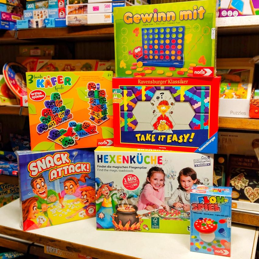 Eine Ausstellung von Brettspielen auf einem Regal, einschließlich 'Gewinn mit', '3 Bündel Kafer', 'Ravensburger Klassiker', 'Take It Easy!', 'Hexenkuche', 'Snack Attack!' und 'Floh-Spiel'. Dahinter befinden sich weitere Spiele und Spielzeug auf den Regalen.