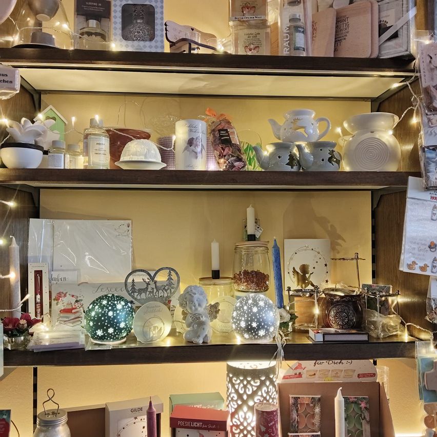 Ein Holzregal gefüllt mit Dekorationsartikeln, einschließlich Kerzen, Figuren und Ornamenten. Verschiedene Artikel sind von Lichterketten beleuchtet, und einige Artikel haben ein Weihnachtsthema.