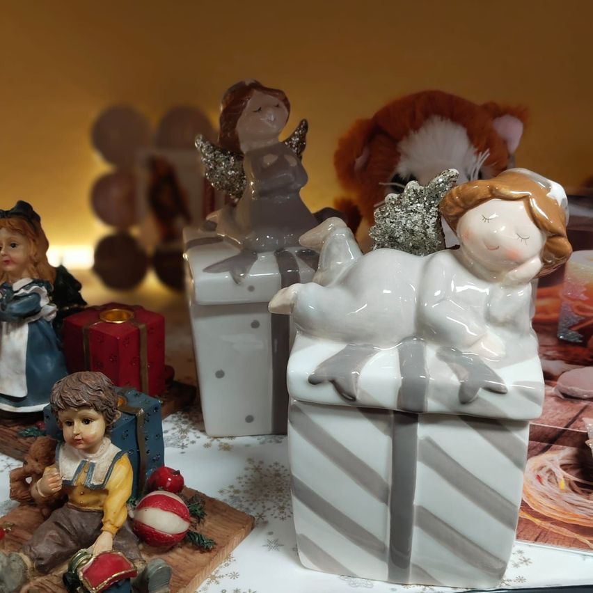 Eine Sammlung von Keramikfiguren, darunter Engel, ein Junge und ein Mädchen, auf einem Tisch mit Geschenken und Spielzeug ausgestellt. Dahinter ein verschwommener Blick auf einen Teddybären und Luftballons.