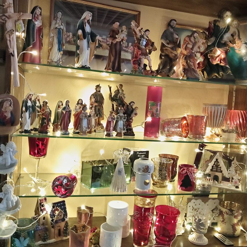 Ein Vitrinenschrank zeigt verschiedene religiöse Figuren, darunter Heilige und Engel, die mit Lichtern geschmückt sind. Die Regale enthalten auch dekorative Kerzen, Tassen und andere Gegenstände. Darüber hängt ein gerahmtes Bild mit einer religiösen Szene mit Figuren.