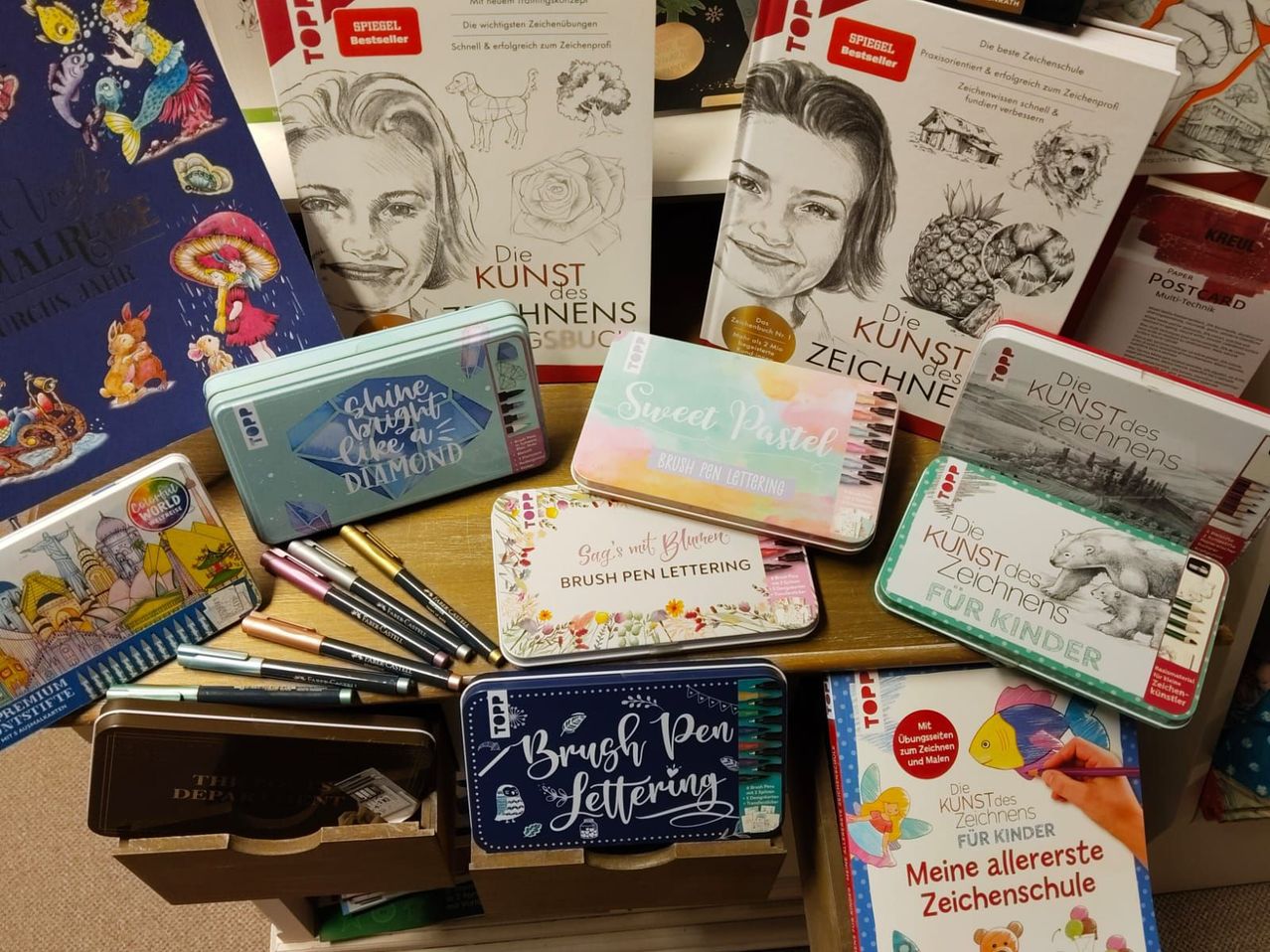 Ein Holztisch zeigt verschiedene Zeichensets und Bücher. Links ein Set mit dem Titel 'Shine Bright Like a Diamond' und ein Set mit 'Sweet Pastel'. In der Mitte zwei 'Brush Pen Lettering'-Sets. Rechts Bücher mit den Titeln 'Die Kunst des Zeichnens' und 'Meine allererste Zeichenschule'.