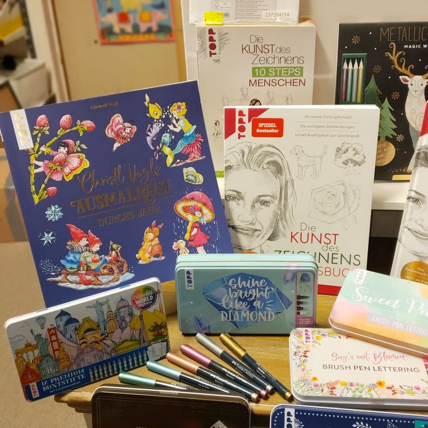 Eine Sammlung von Malbüchern, Pinseln und Zeichenutensilien auf einem Holztisch. Titel beinhalten 'Aussie Magic', 'Kunst des Zeichnens' und 'Metallic Magic'. Verschiedene Buntstifte und Stifte sind in der Nähe angeordnet.