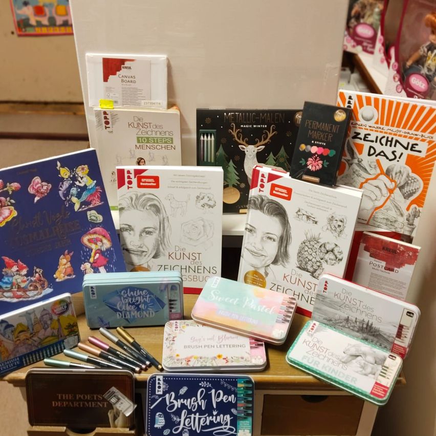 Eine Ausstellung verschiedener Zeichenbücher und Werkzeuge. Titel beinhalten 'Kunst des Zeichnens,' 'Brush Pen Lettering' und 'Permanent Marker.' Bunte Dosen mit Stiften und Pinseln sind ebenfalls vorhanden.